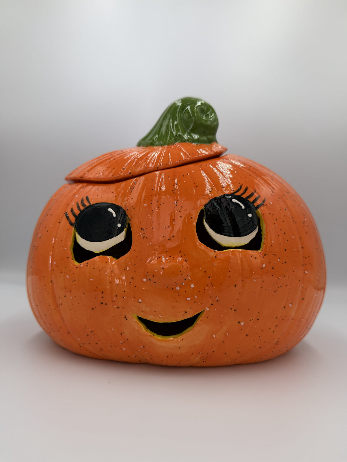 Orange speckled ceramic jack-o'-lantern with big lashed eyes, smile, and green stem lid