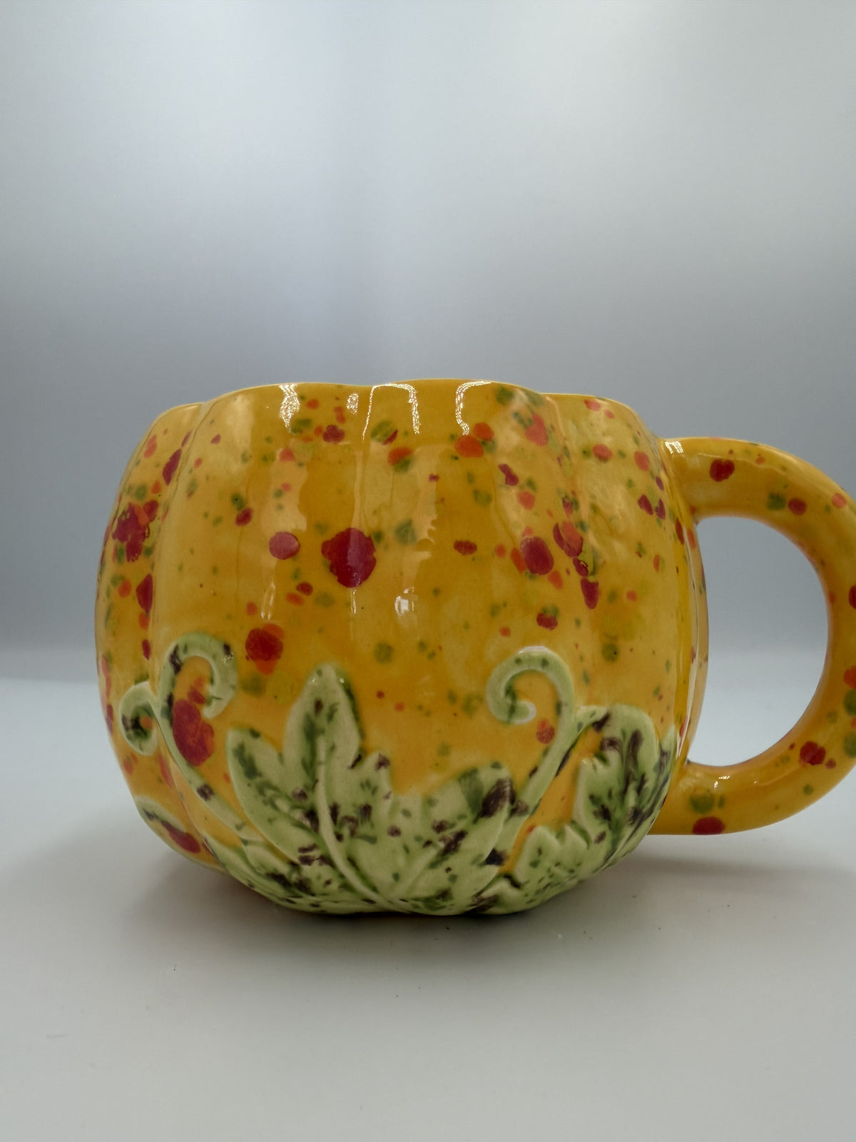 Yellow ceramic pumpkin mug with red, orange, and green speckles and sculpted green leaves at the base